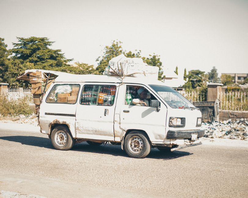 Old white van loaded with goods driving on a sunny street, capturing rustic charm.