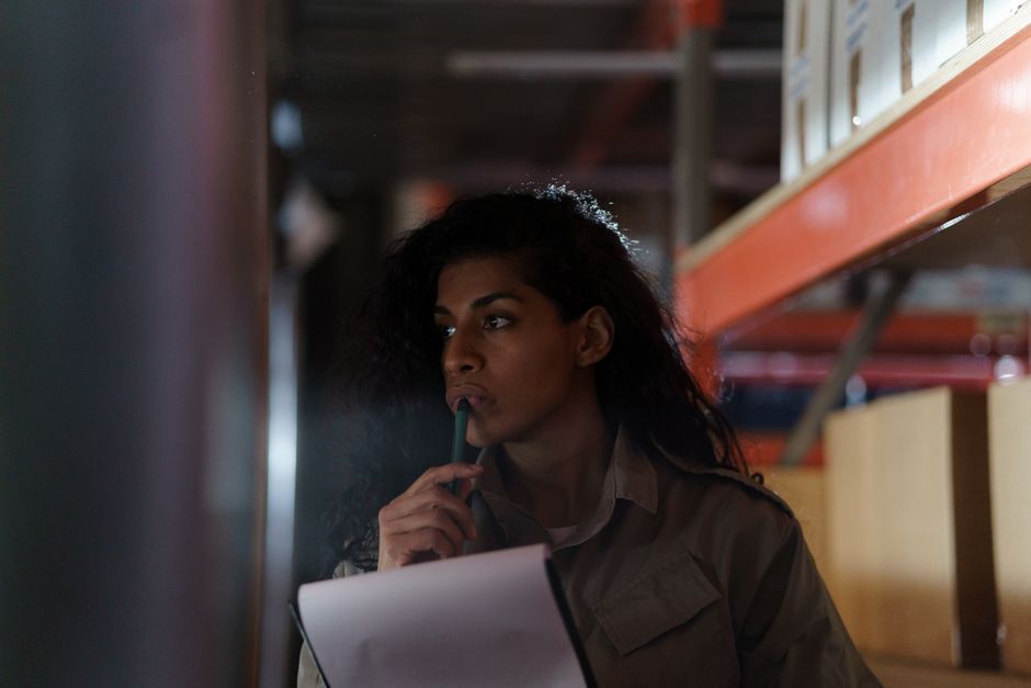 A woman in a warehouse contemplatively checking inventory under low light conditions.