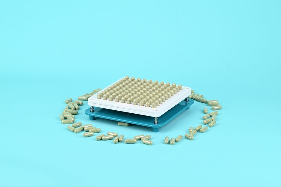 Manual capsule filling tray surrounded by herbal supplements on a blue backdrop, ideal for health and wellness themes.