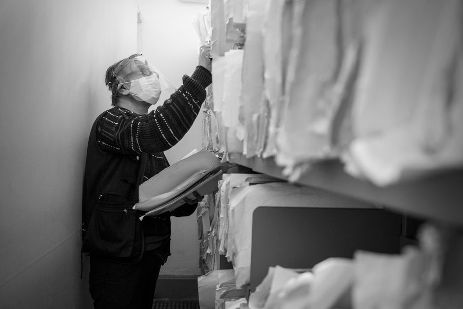 Black and white image of a masked elderly man organizing records in an office aisle.