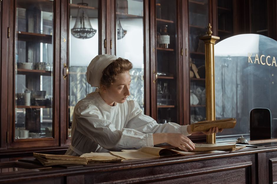 A woman chemist in a vintage pharmacy reads a book behind the counter.
