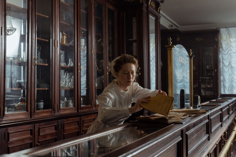 A woman in a retro pharmacy setting, organizing papers at the counter.