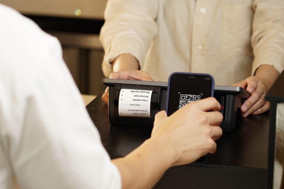 Two individuals engaged in a contactless payment using a QR code scanner at a retail counter.