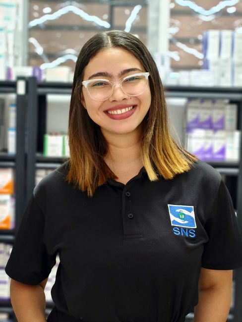 Smiling woman in a pharmacy setting wearing a black SNS uniform, indoors.