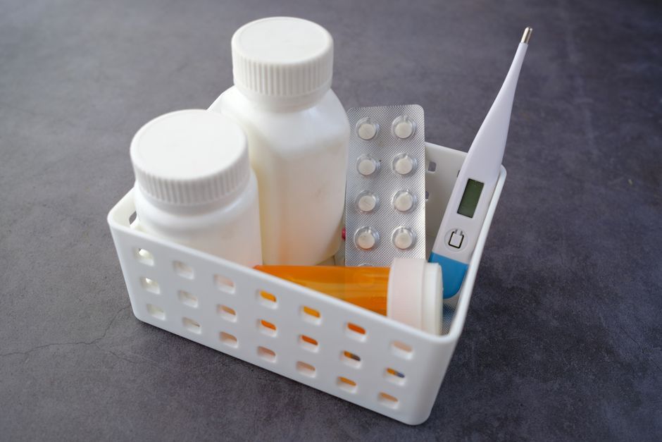 Healthcare basket with medication bottles, pills, and thermometer on a dark surface.