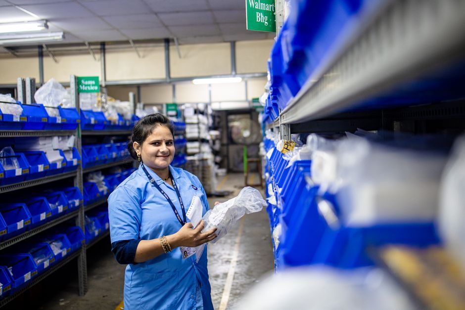 Smiling woman in a warehouse organizing inventory on shelves with blue bins.